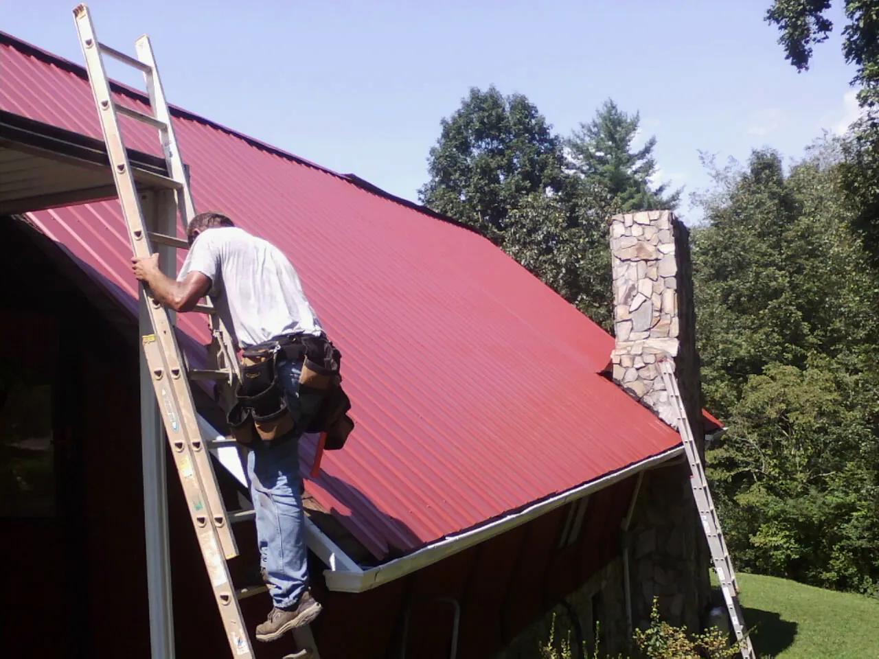 Roofer on a ladder beside a red metal roof and stone chimney — Soffit & Fascia Repair by Roofing Brothers in Las Animas